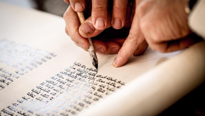 Hands of a scribe holding a quill and touching it to.a Torah scroll.