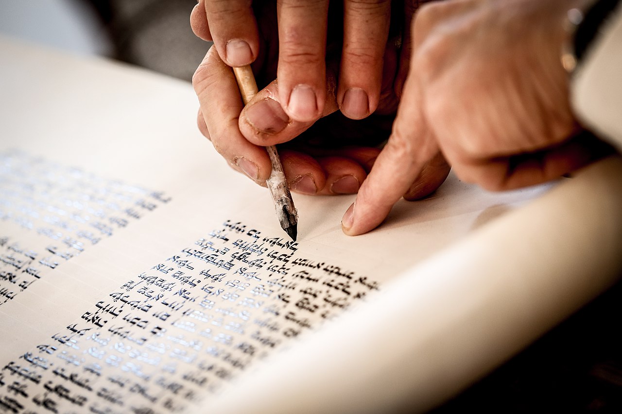 Hands of a scribe holding a quill and touching it to.a Torah scroll.