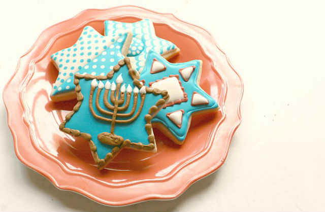 Four frosted sugar cookies are on a plate. The cookies are each shaped like a Star of David and decorated with Hanukkah motifs, like a menorah. The cookies are all blue and white.