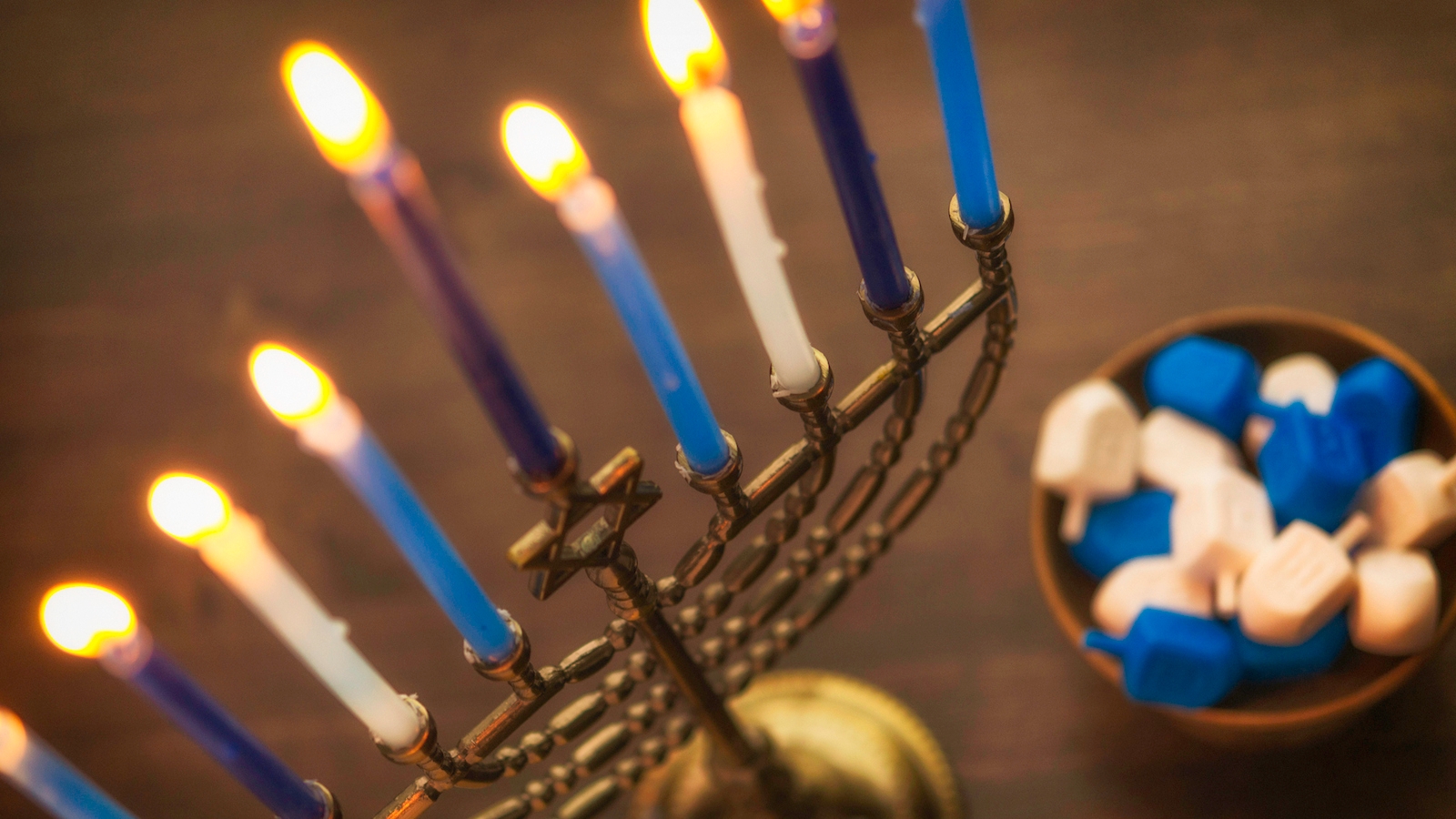 A bronze Hanukkah menorah with nine blue and white candles that are all lit. One of the candles, the shamash, is higher than the others. There are dreidels next to the menorah.