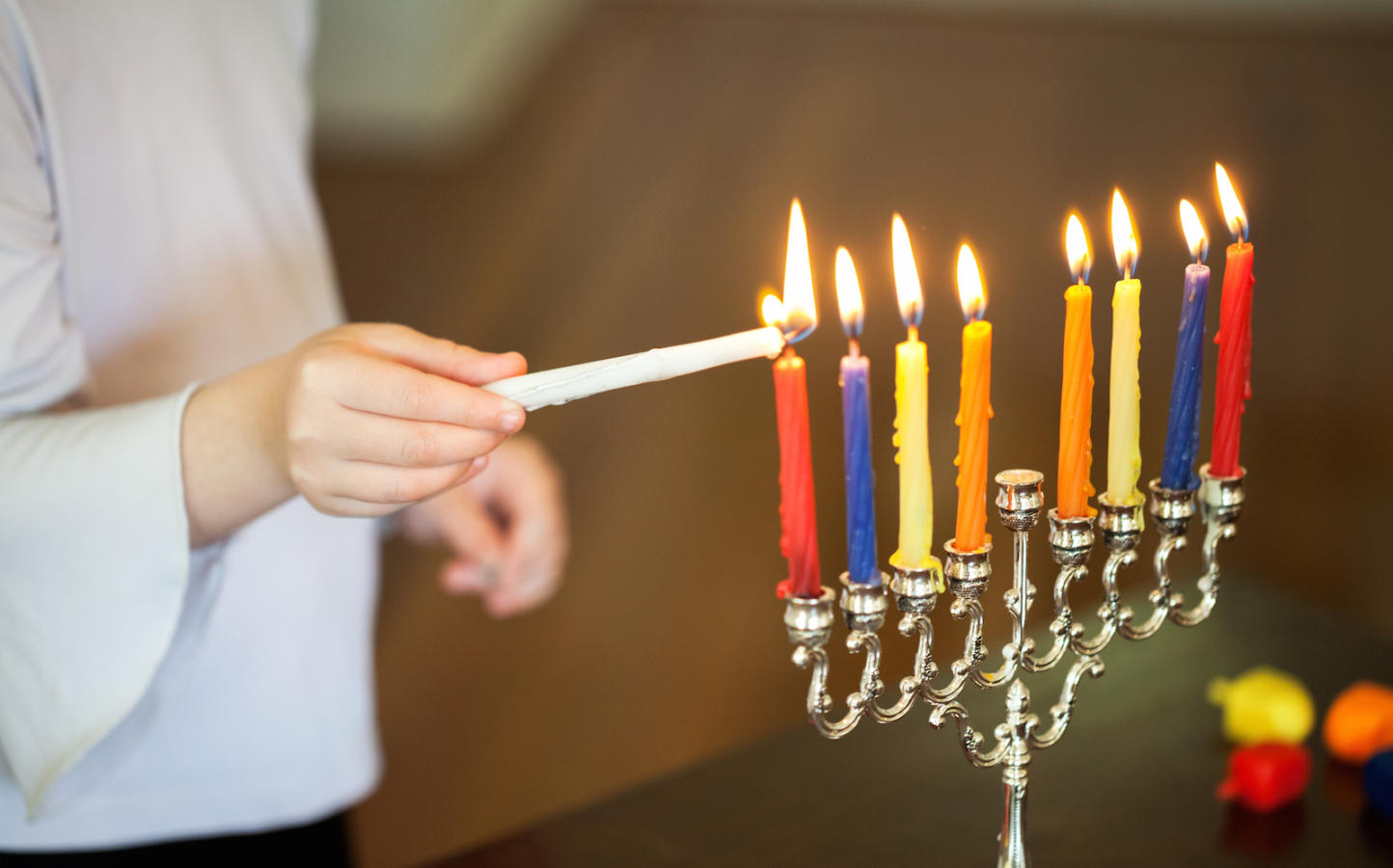 A child uses the shamash to light the Hanukkah candles. The candles are inside a silver menorah.