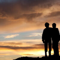 Two happy young boys on a hill at sunset. Back view. Children are elementary aged and have their arms around each other in embrace. Unrecognizable models. Themes in the image include best friends, love, relationships, siblings, brothers, bonding, care, comfort, talking, outside, nature, recreation, leisure, watching the sunset, beauty, and family.