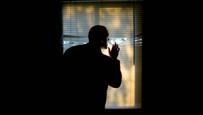 A man peeking outdoors through the blinds of a darkened room