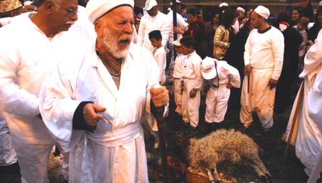 Man in a robe with an intense look on his face next to a slaughtered sheep with a crowd of peple.