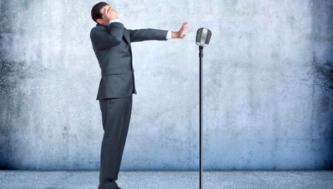 A businessman covers his eyes and holds out an outstreched arm as he steps up to a microphone on a stand, exhibiting his fear of public speaking.